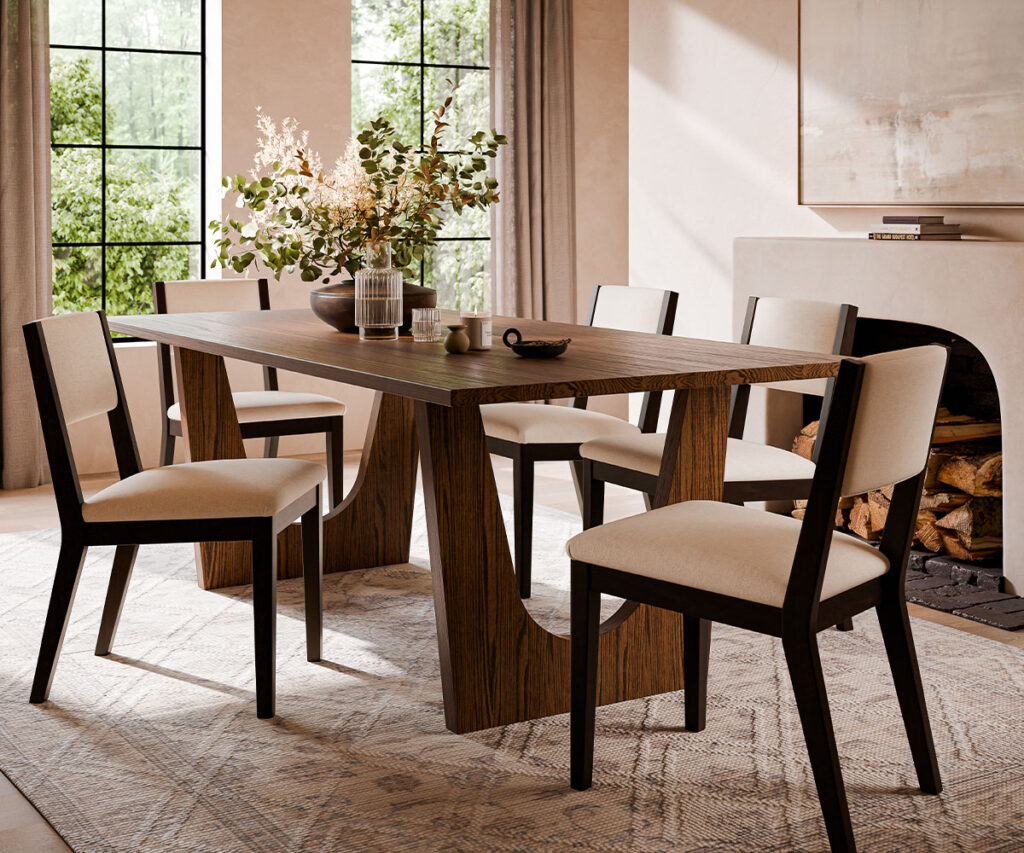 A modern heirloom furniture investment featuring a sleek wooden dining table with sculptural legs and six upholstered chairs in a neutral-toned dining space. Large windows bring in natural light, complementing the warm wood tones, soft rug, and fireplace, creating a timeless yet contemporary setting built for lasting elegance.