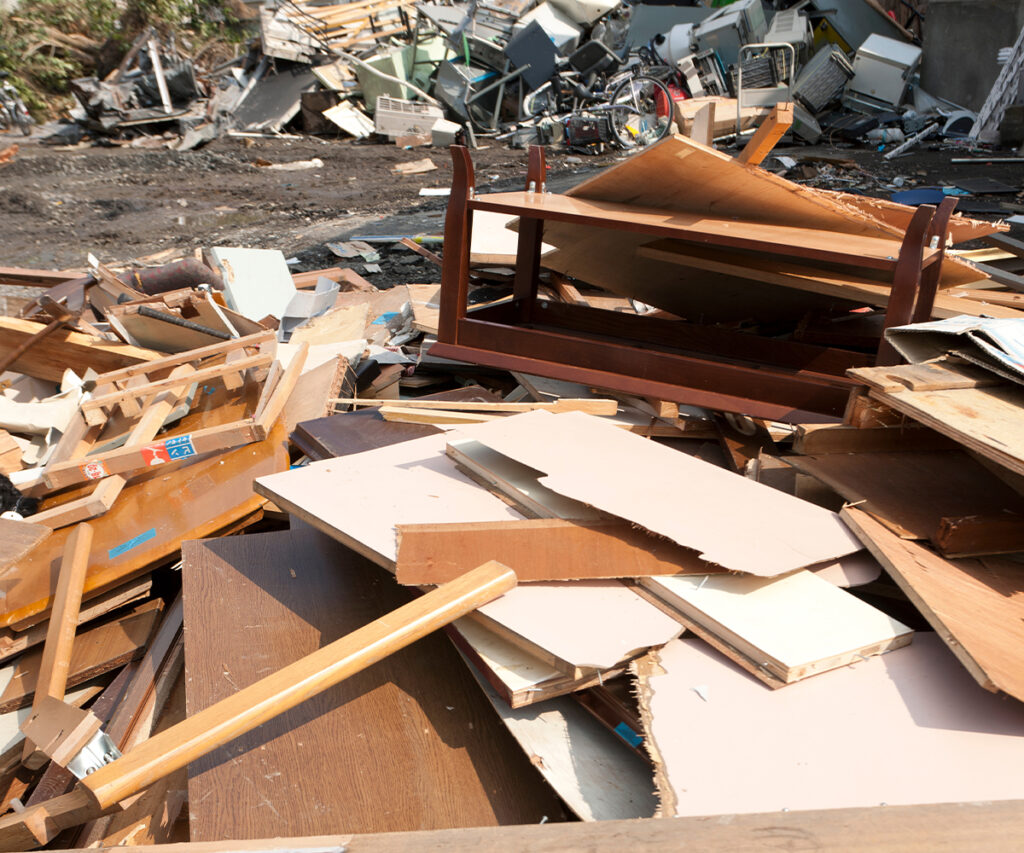 A pile of broken and discarded furniture, including shattered particleboard, splintered wood, and an overturned table, sits in a landfill filled with waste and debris. This image contrasts the fleeting nature of mass-produced furniture with the lasting value of an heirloom furniture investment, which is built to endure generations rather than end up as waste.