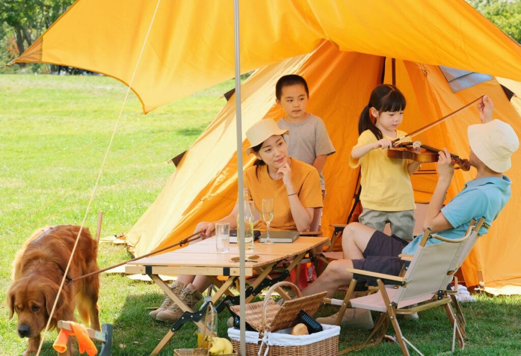 Family camping near orange tent as child plays violin.