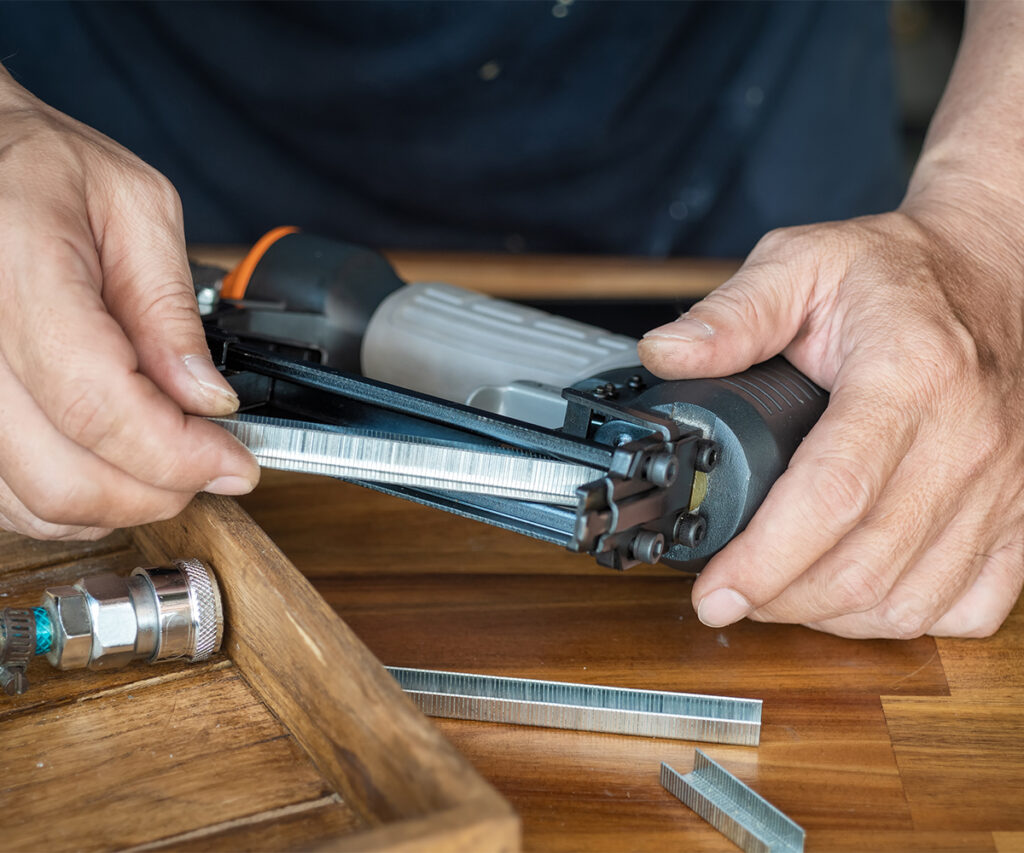 Hands using pneumatic stapler on wood frame, showing fasteners used in furniture assembly.